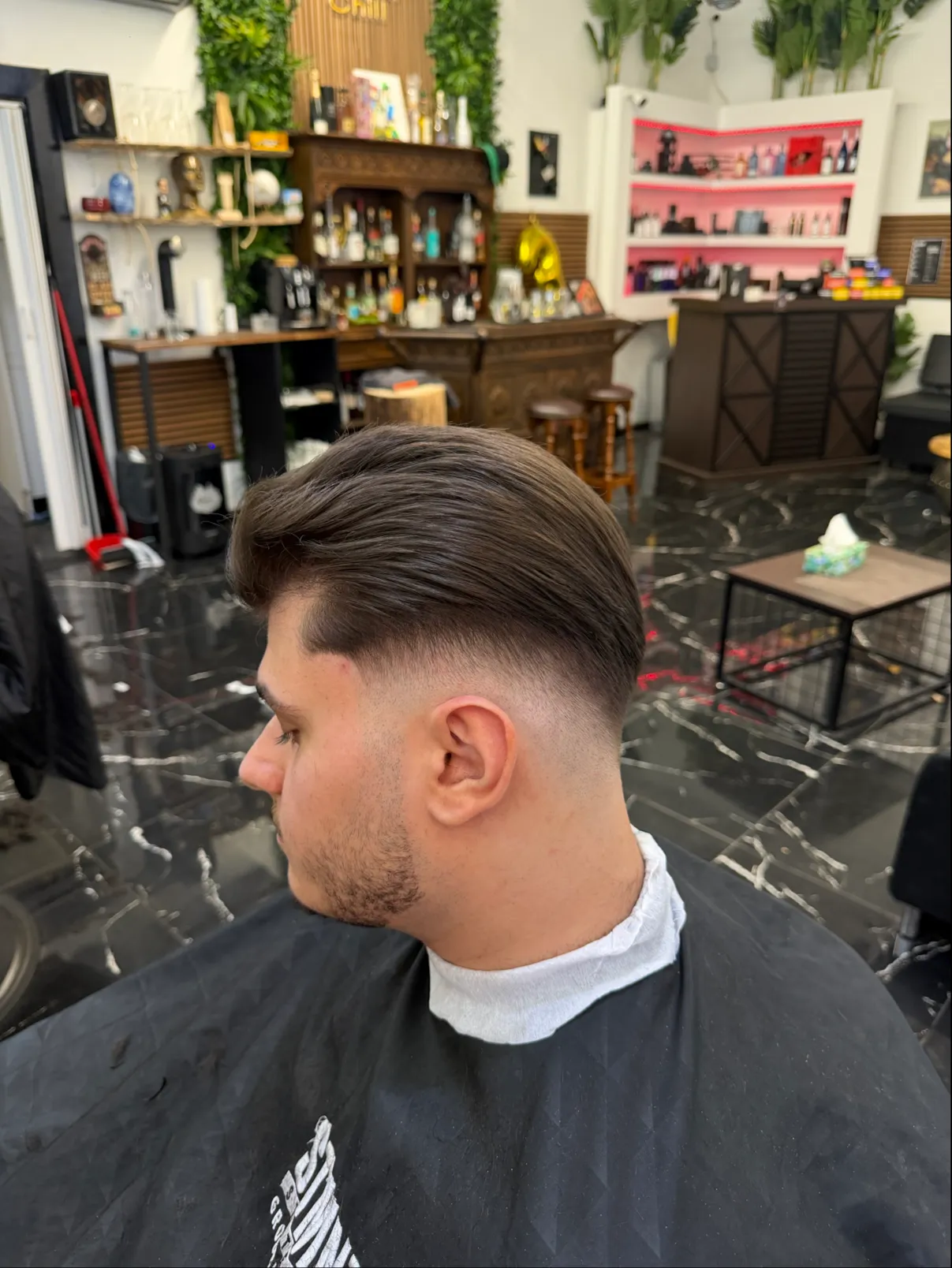 Frischer Low Fade mit Slick Back, im Hintergrund der Laden mit schwarzem Marmorboden und rosa beleuchteten Regalen